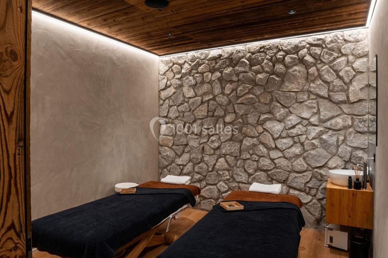 Salle de soins avec deux tables de massage, murs en pierre, éclairage tamisé et ambiance chaleureuse.