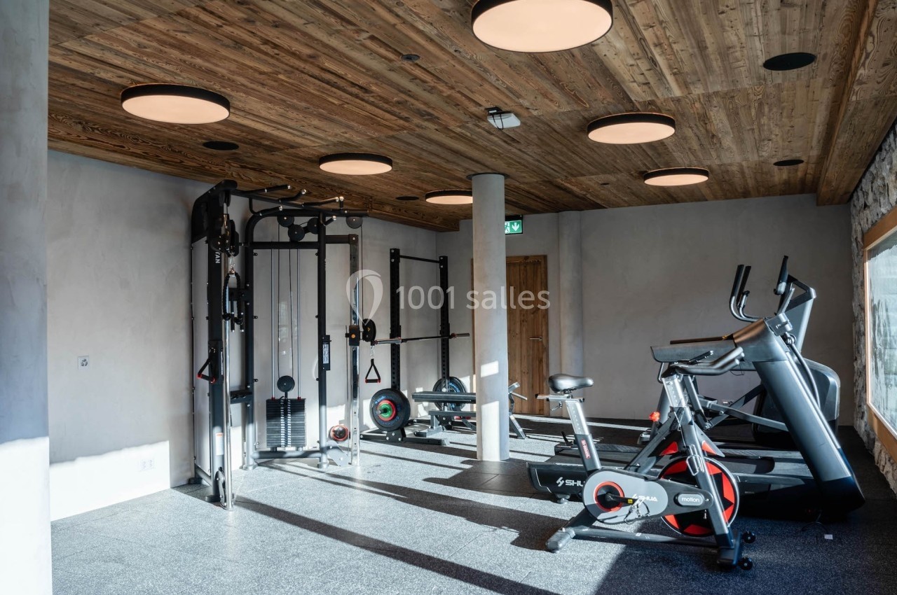 Salle de sport lumineuse avec équipements de musculation, tapis de course et vélo d'entraînement, plafond en bois.