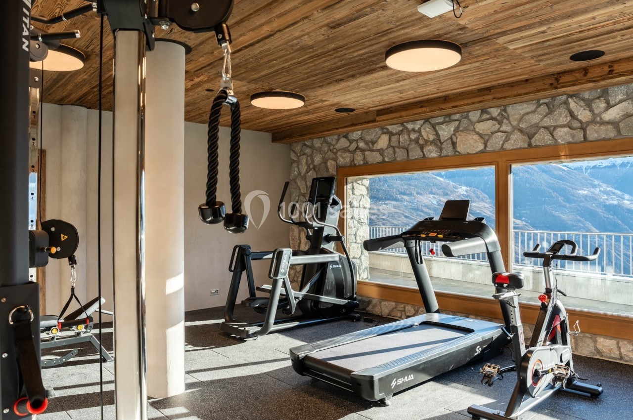 Salle de sport lumineuse avec tapis de course, vélo d'appartement et équipements, vue sur les montagnes à travers de grandes…