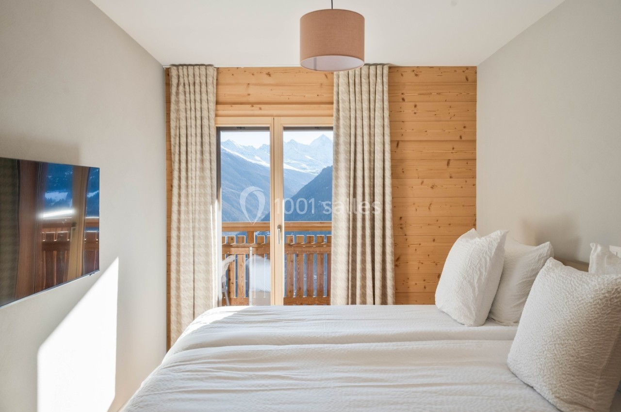 Chambre lumineuse avec lit double, murs en bois, rideaux beiges et vue sur des montagnes enneigées par une baie vitrée.