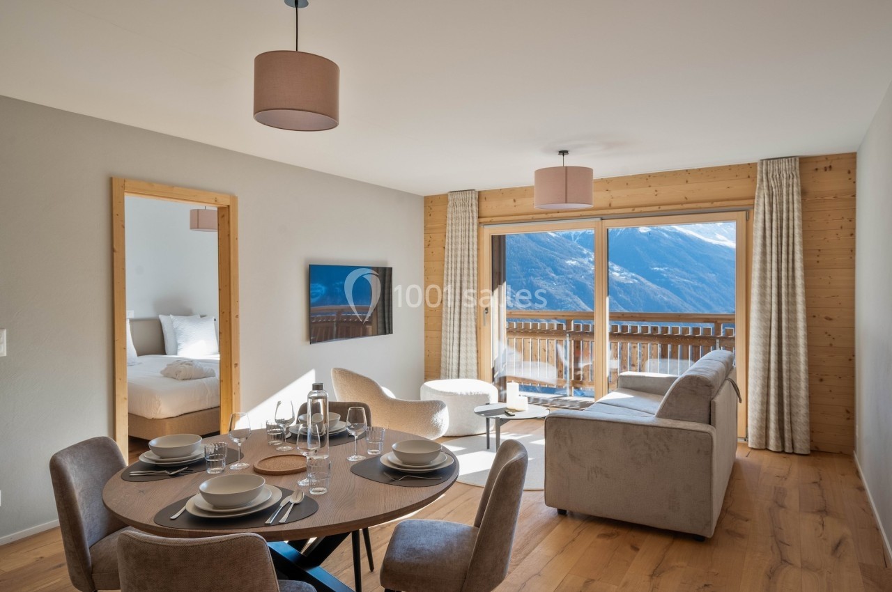 Salon lumineux avec canapé, table dressée, télévision murale et grande baie vitrée donnant sur une vue montagne.