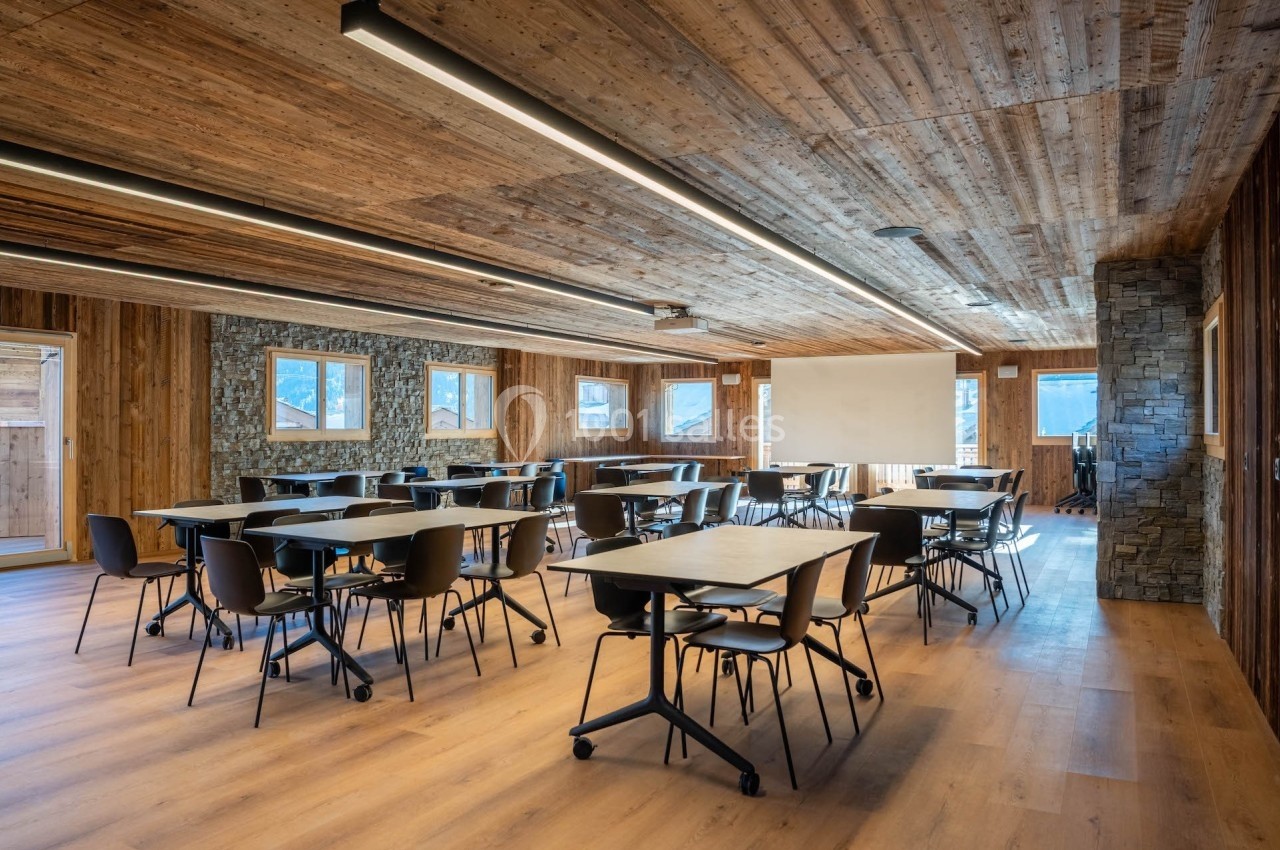 Salle de réunion lumineuse avec tables et chaises modernes, murs en bois et pierre, et écran de projection.