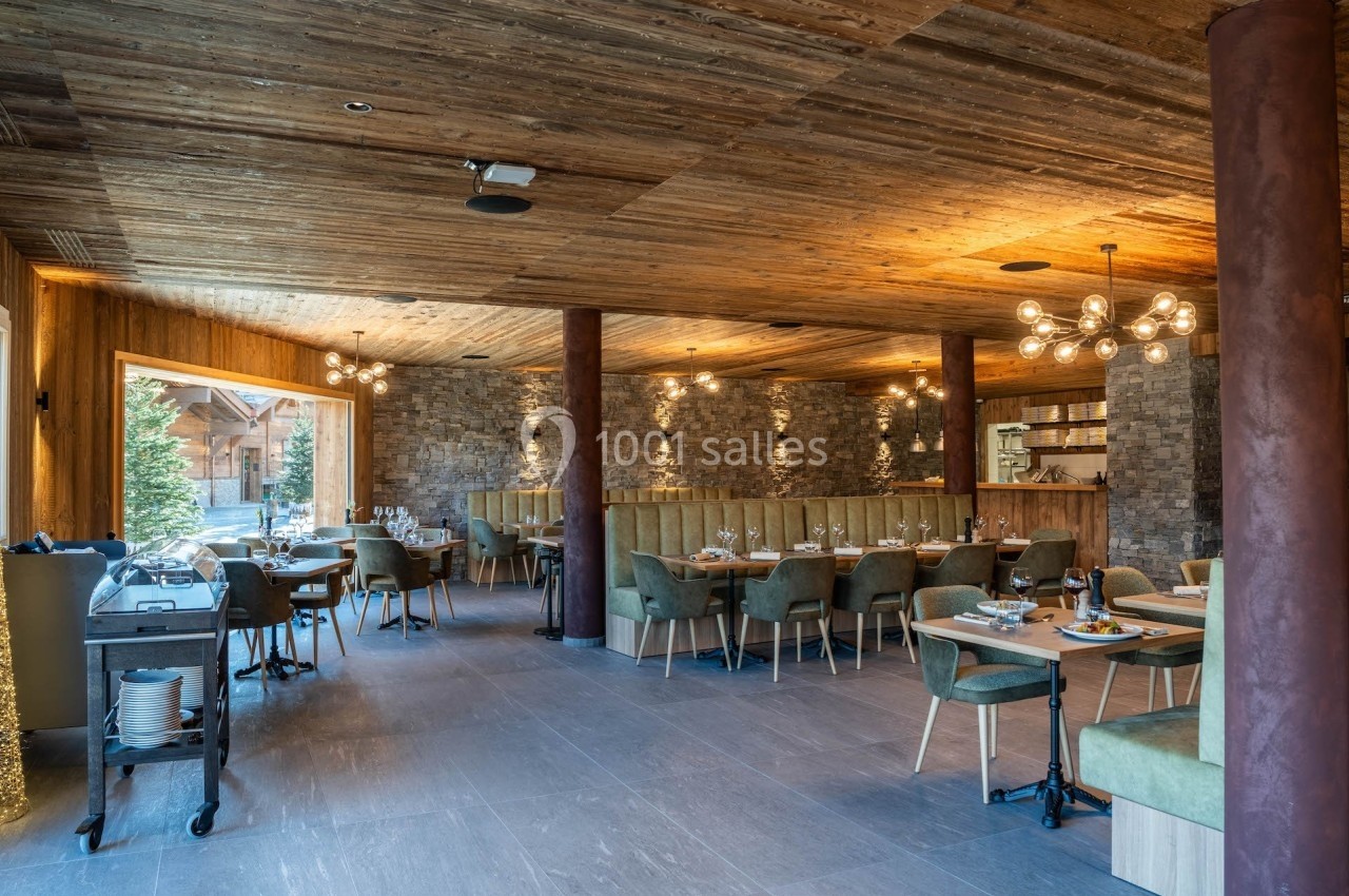 Salle de restaurant élégante avec tables dressées, chaises vertes, murs en pierre et plafond en bois éclairé.