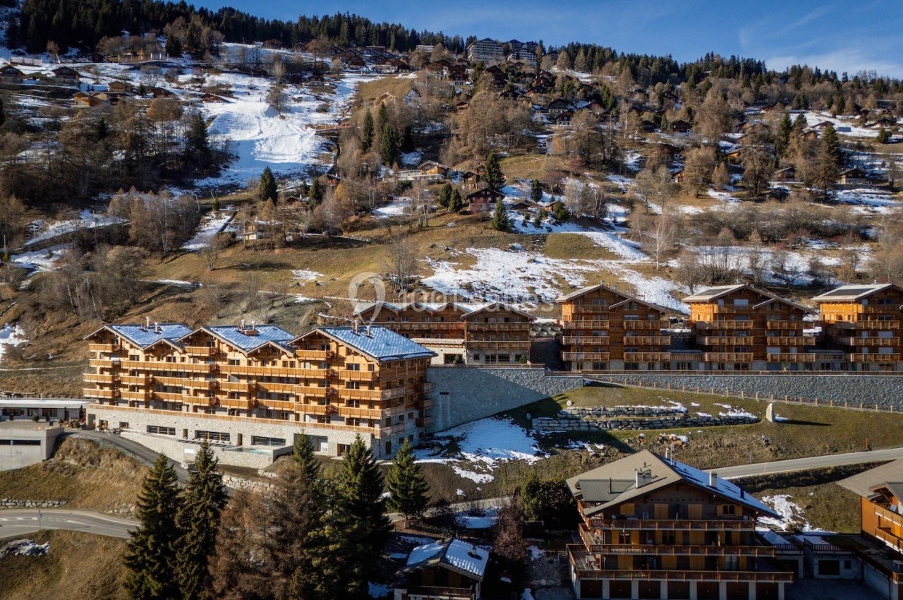 Vue aérienne de chalets en bois sur une colline enneigée, entourés de végétation et de routes sinueuses.