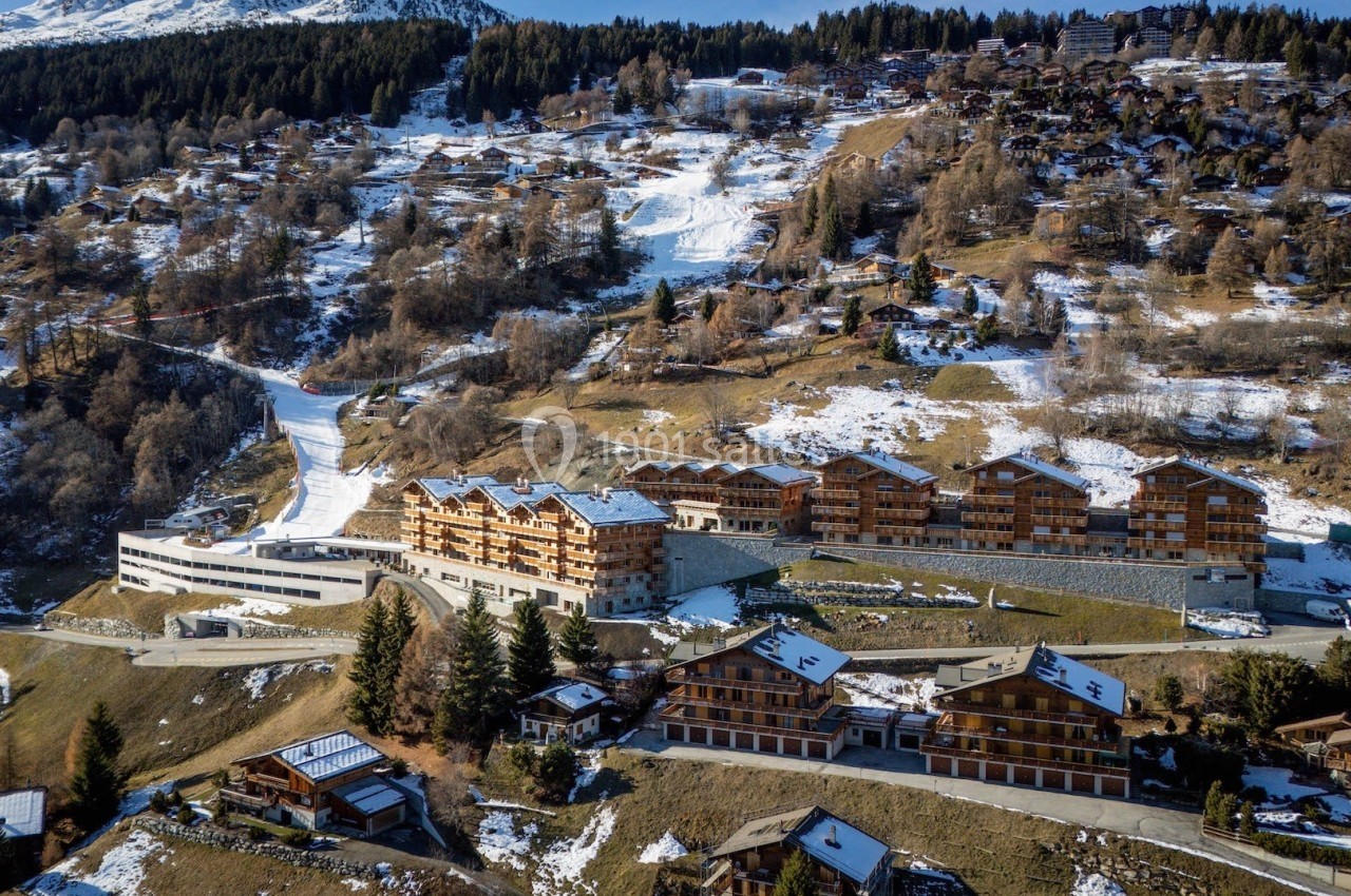 Vue aérienne d'une station de montagne avec chalets en bois, pistes de ski et forêt en arrière-plan.