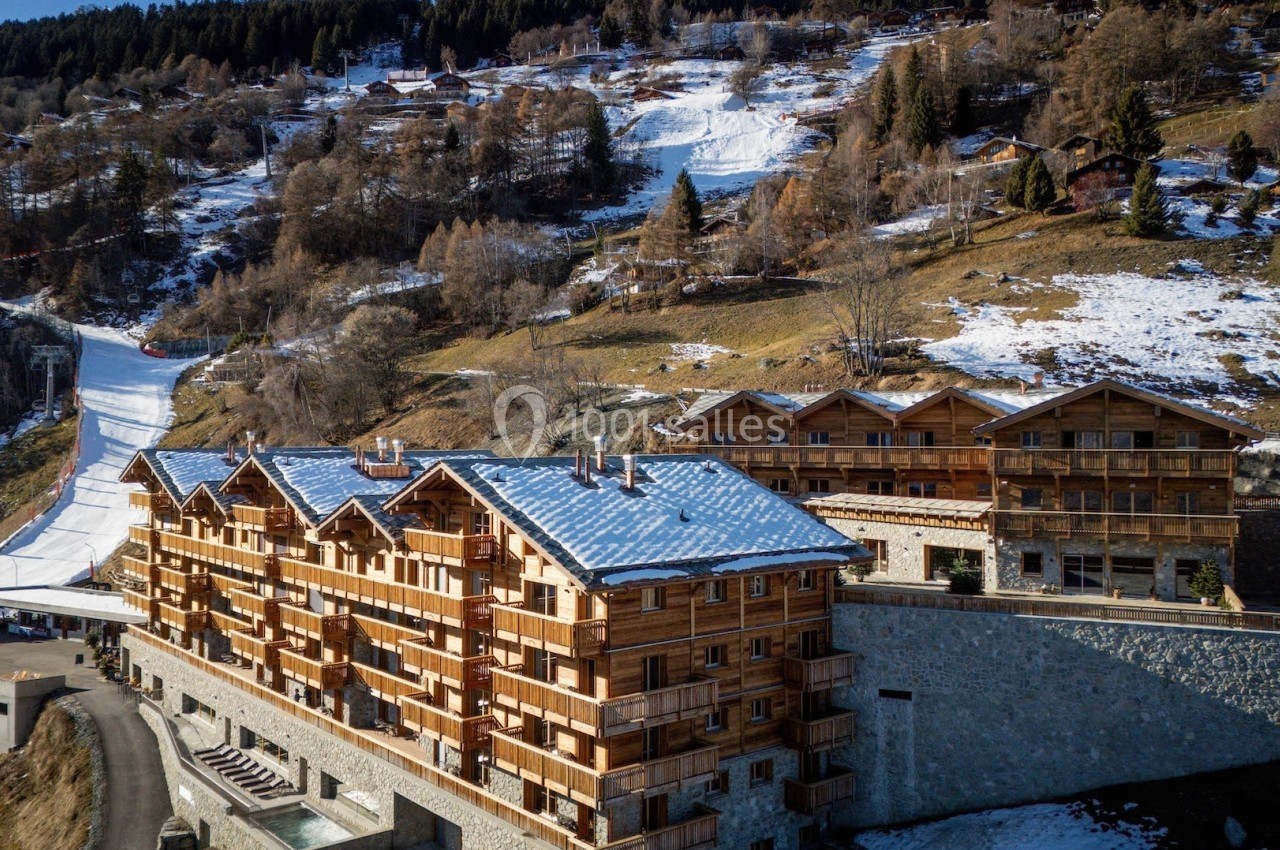 Vue aérienne d'un complexe de chalets en bois près d'une piste de ski, entouré de montagnes partiellement enneigées.