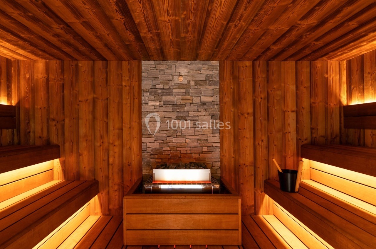 Intérieur d'un sauna en bois avec bancs éclairés, un mur en pierre et un seau en bois posé sur le côté.