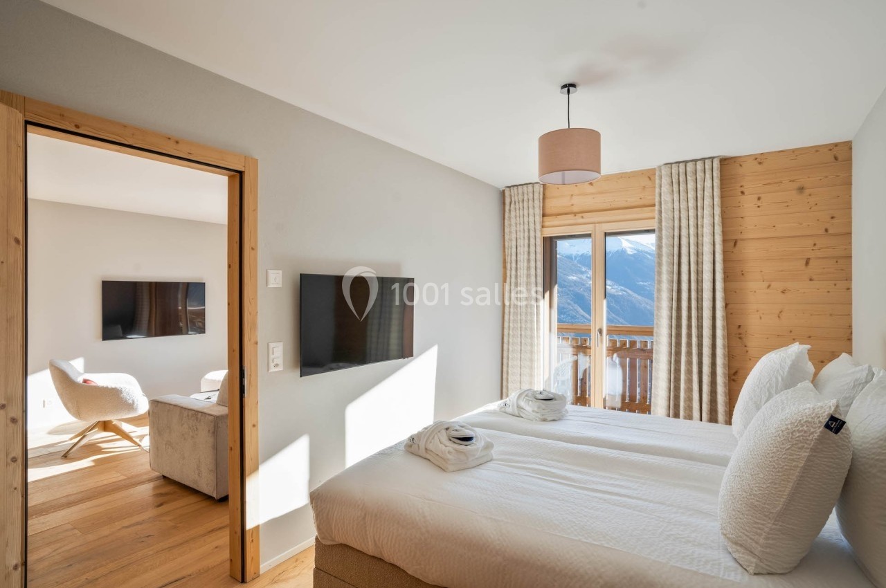 Chambre lumineuse avec deux lits simples, télévision murale et vue sur les montagnes depuis une porte-fenêtre.