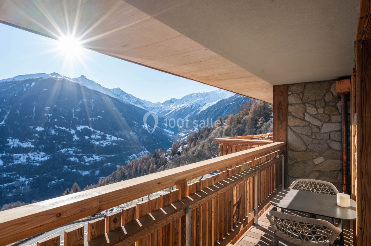 Vue depuis un balcon en bois sur une vallée enneigée et des montagnes sous un ciel ensoleillé.