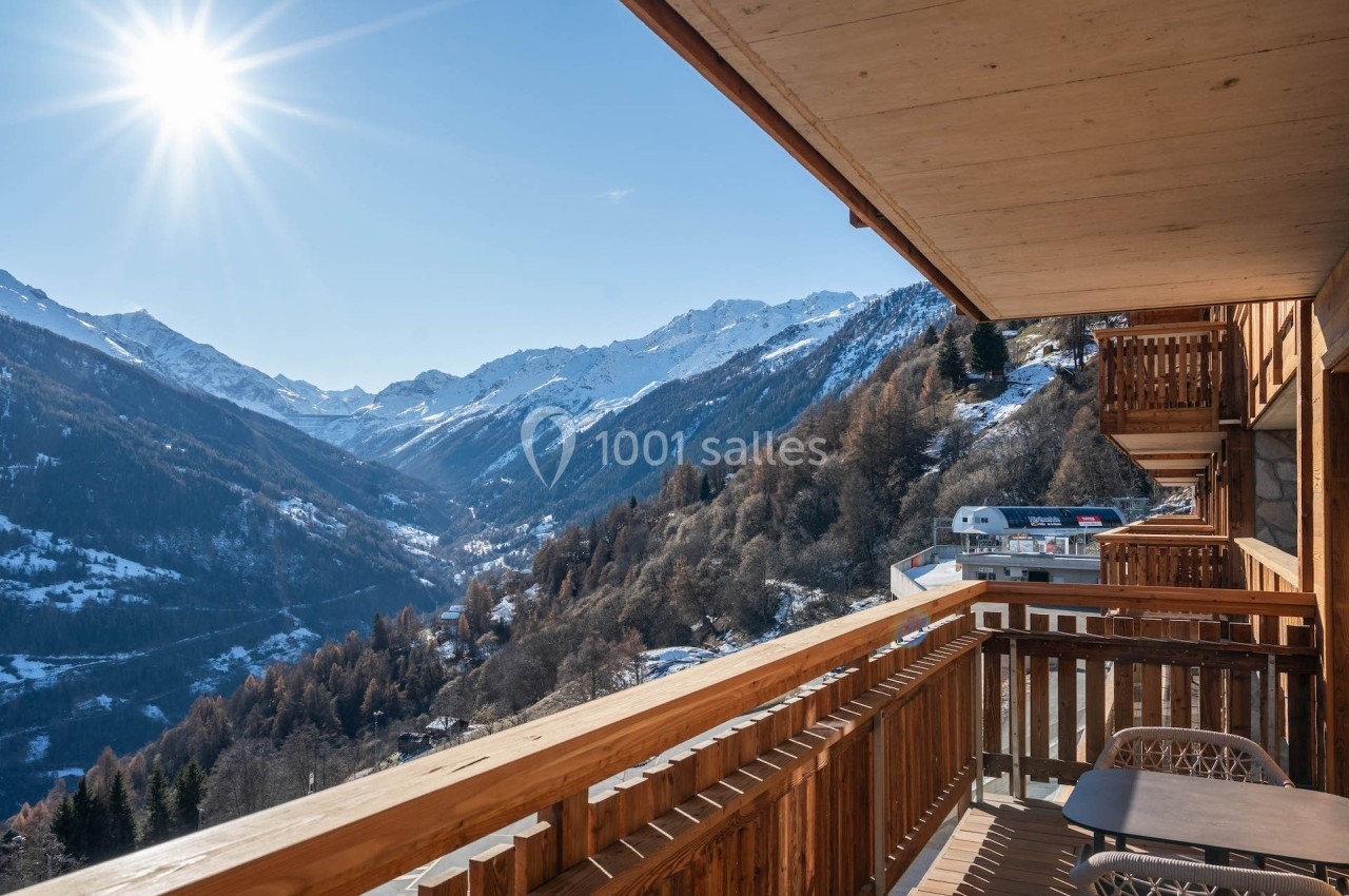 Vue depuis un balcon en bois sur des montagnes enneigées sous un ciel bleu ensoleillé.