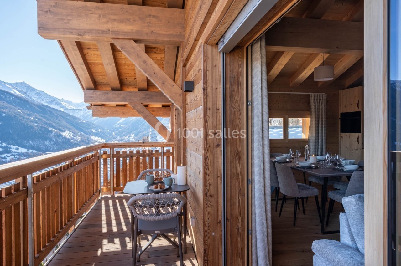Balcon en bois avec vue sur des montagnes enneigées, jouxtant une salle à manger lumineuse dans un chalet.