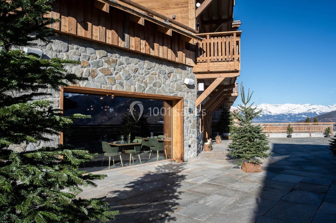 Façade en pierre et bois d'un chalet avec terrasse, sapins et vue sur des montagnes enneigées sous un ciel bleu.