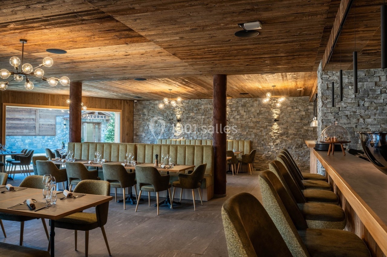 Salle de restaurant chaleureuse avec tables dressées, banquettes, chaises rembourrées et murs en pierre apparente.