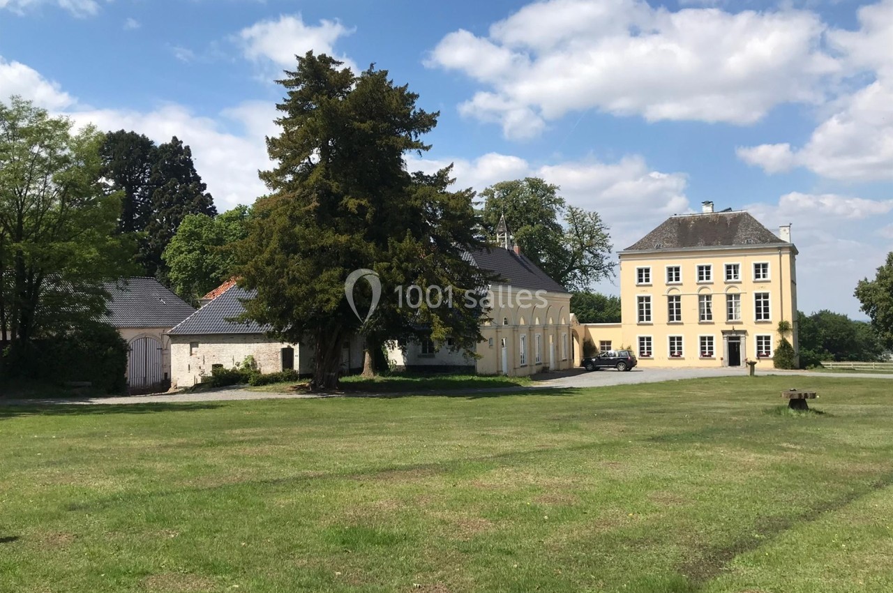 Bâtiments anciens entourés de verdure, comprenant une maison principale jaune et des annexes en pierre sous un ciel dégagé.