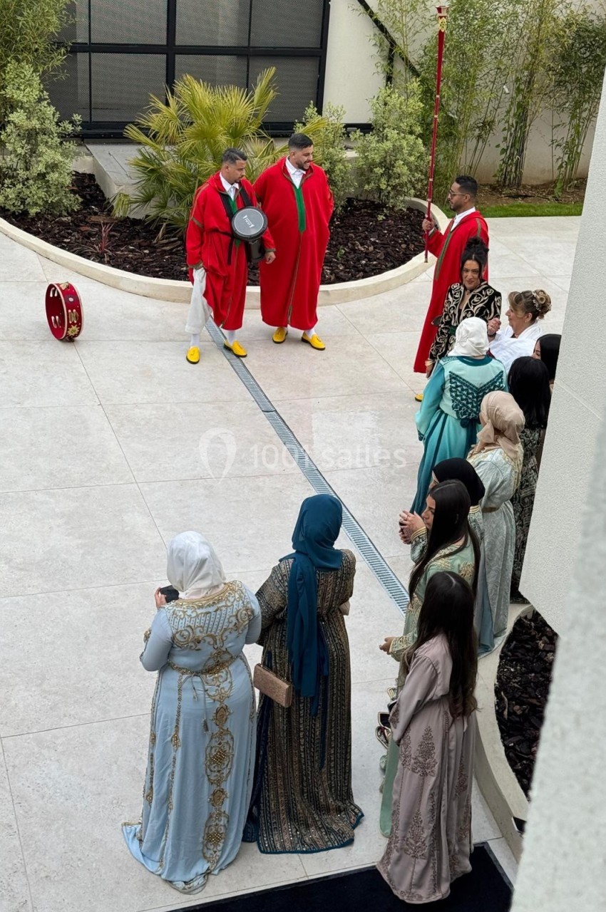 Des musiciens en tenue rouge jouent du tambour devant un groupe de femmes en robes traditionnelles dans une cour extérieure.
