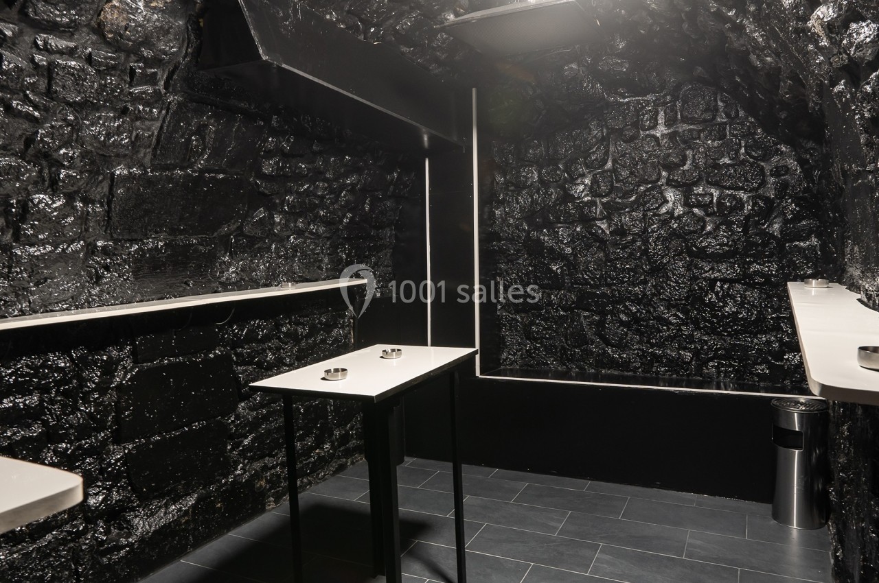 Salle sombre aux murs en pierre noire, équipée de tables hautes blanches et d'un sol en carrelage gris.