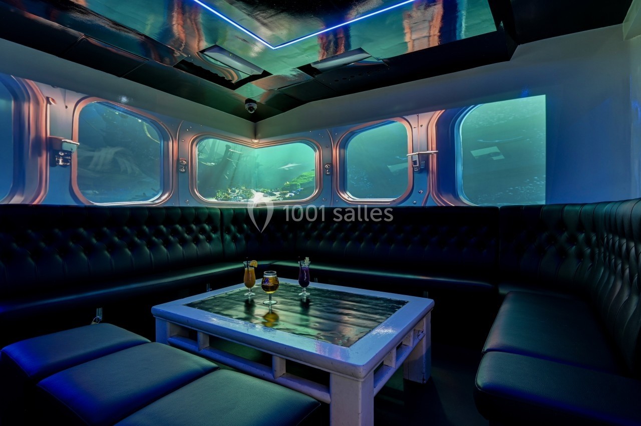Salle lounge sous-marine avec banquettes noires, table basse et grandes fenêtres offrant une vue sur l'océan.