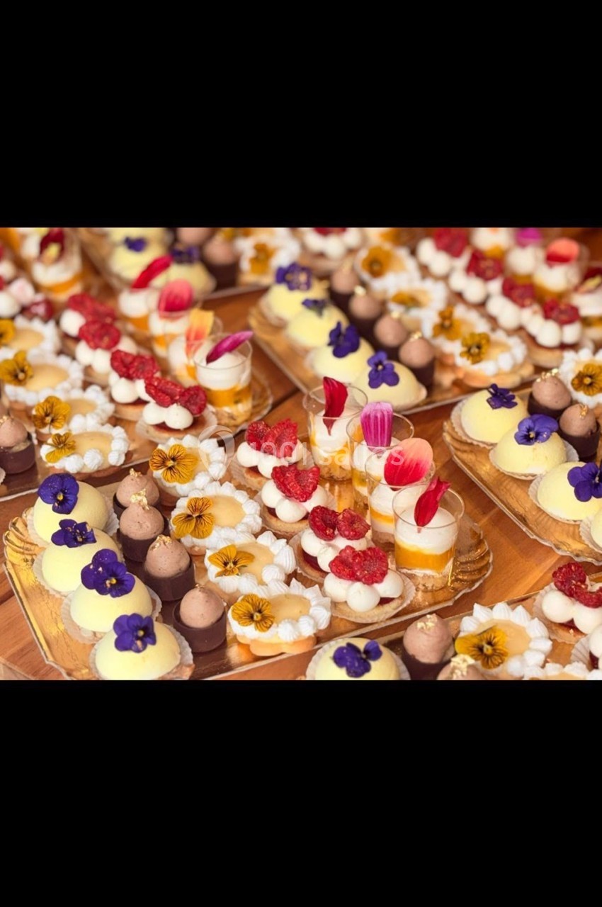Assortiment de pâtisseries décorées de fleurs comestibles, fruits rouges et crèmes colorées sur une table en bois.
