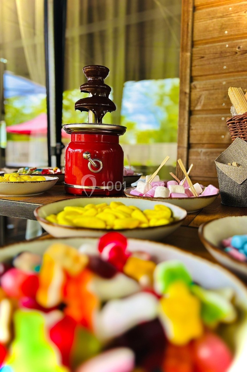 Fontaine à chocolat rouge entourée de bonbons colorés, guimauves et biscuits sur une table en bois.