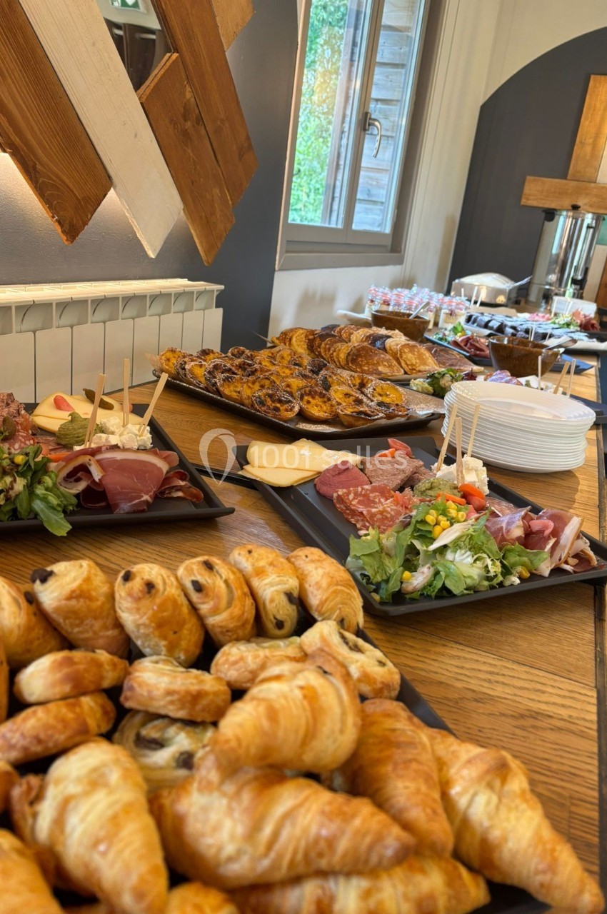 Buffet composé de viennoiseries, charcuteries, salades et desserts, disposé sur une table en bois près d'une fenêtre.