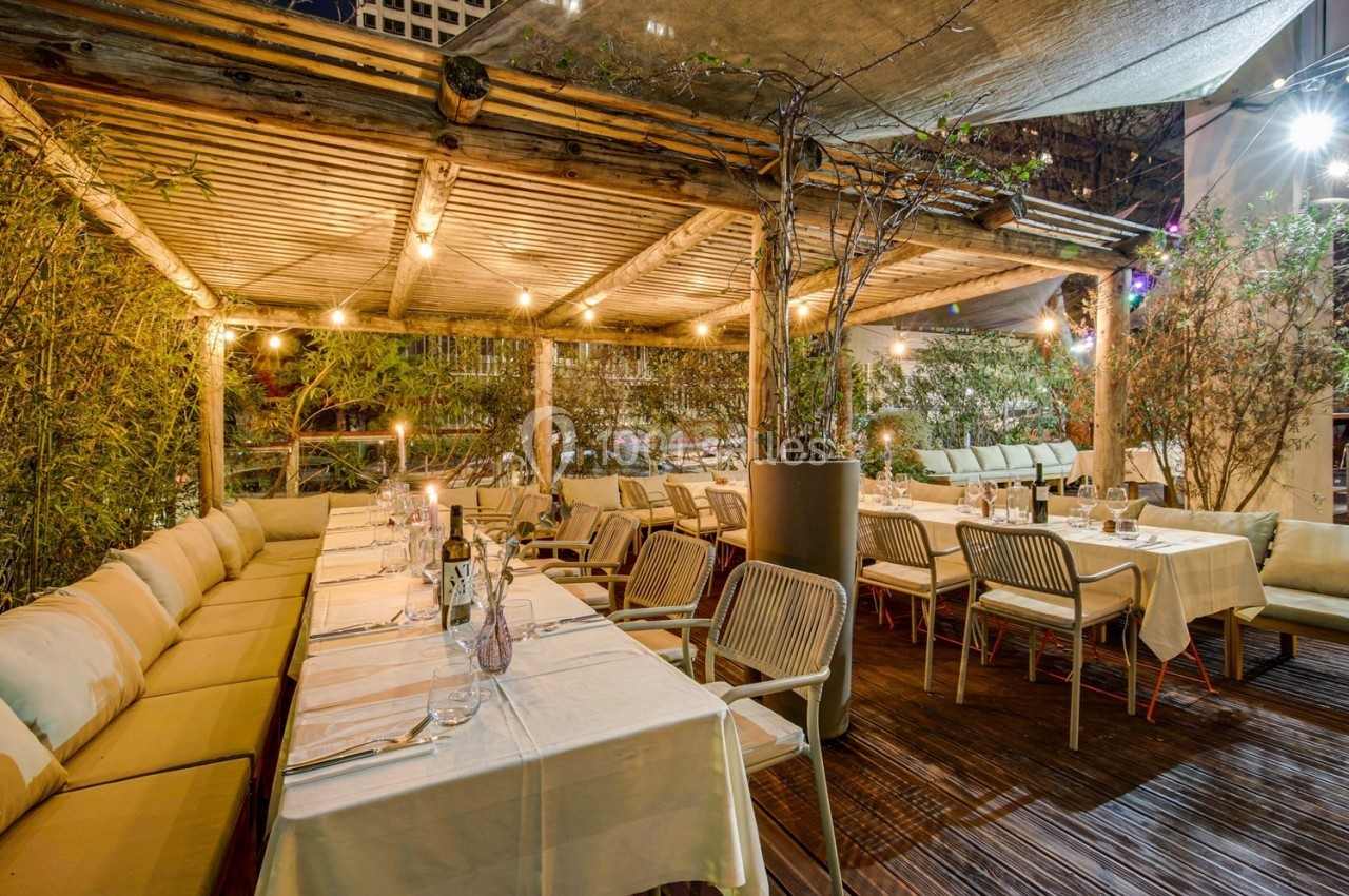 Terrasse extérieure éclairée en soirée avec tables dressées, chaises en bois et végétation environnante.