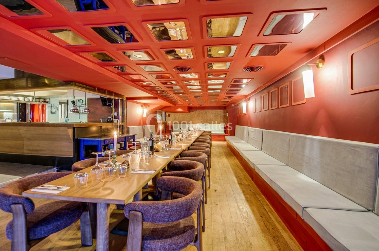 Salle de restaurant avec longues tables en bois, chaises rembourrées, banquettes grises et plafond rouge avec miroirs.