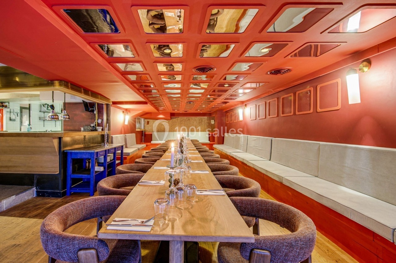 Salle de restaurant avec longues tables en bois, chaises rembourrées, banquettes et plafond rouge à panneaux réfléchissants.