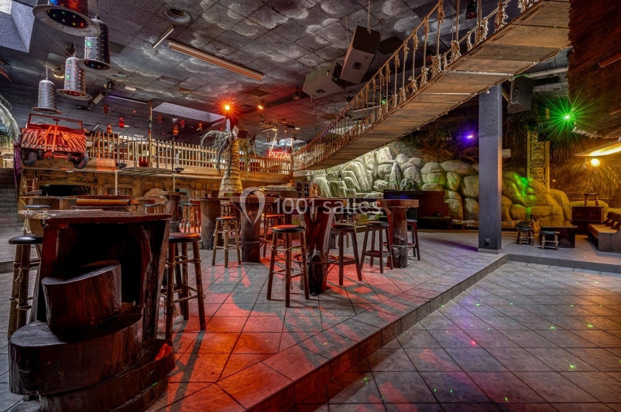 Salle de bar à thème avec pont suspendu, tables en bois et éclairage coloré dans une ambiance tropicale.