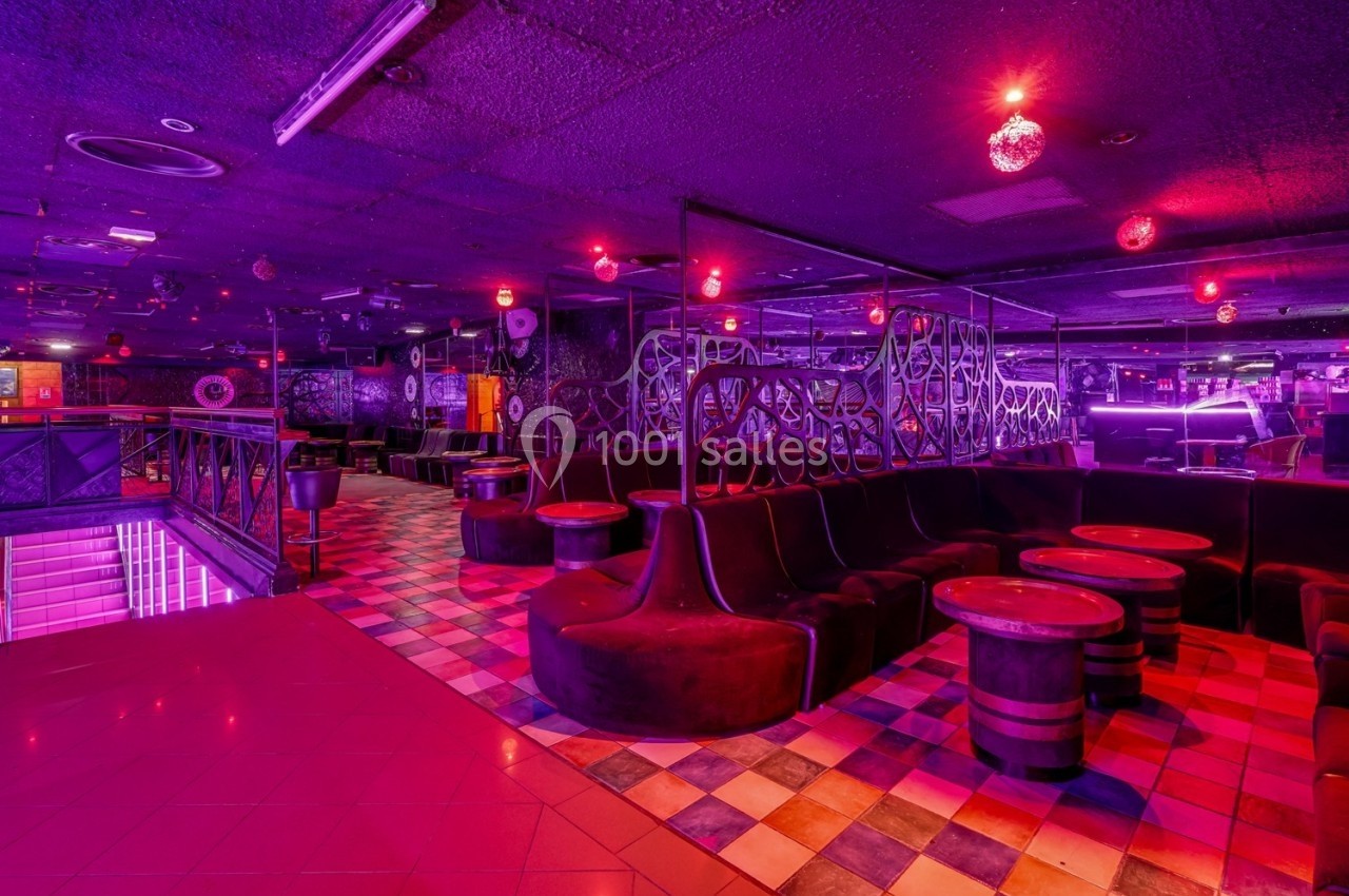 Salle de club nocturne avec éclairage rouge et violet, banquettes noires, tables en tonneaux et sol carrelé.