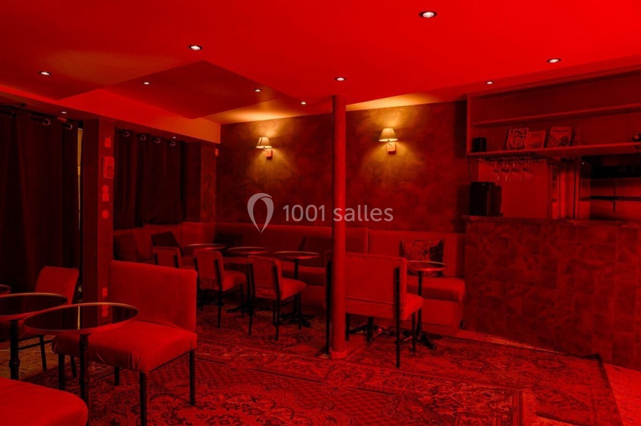 Salle de bar à l'éclairage rouge, avec tables, chaises et comptoir en arrière-plan.