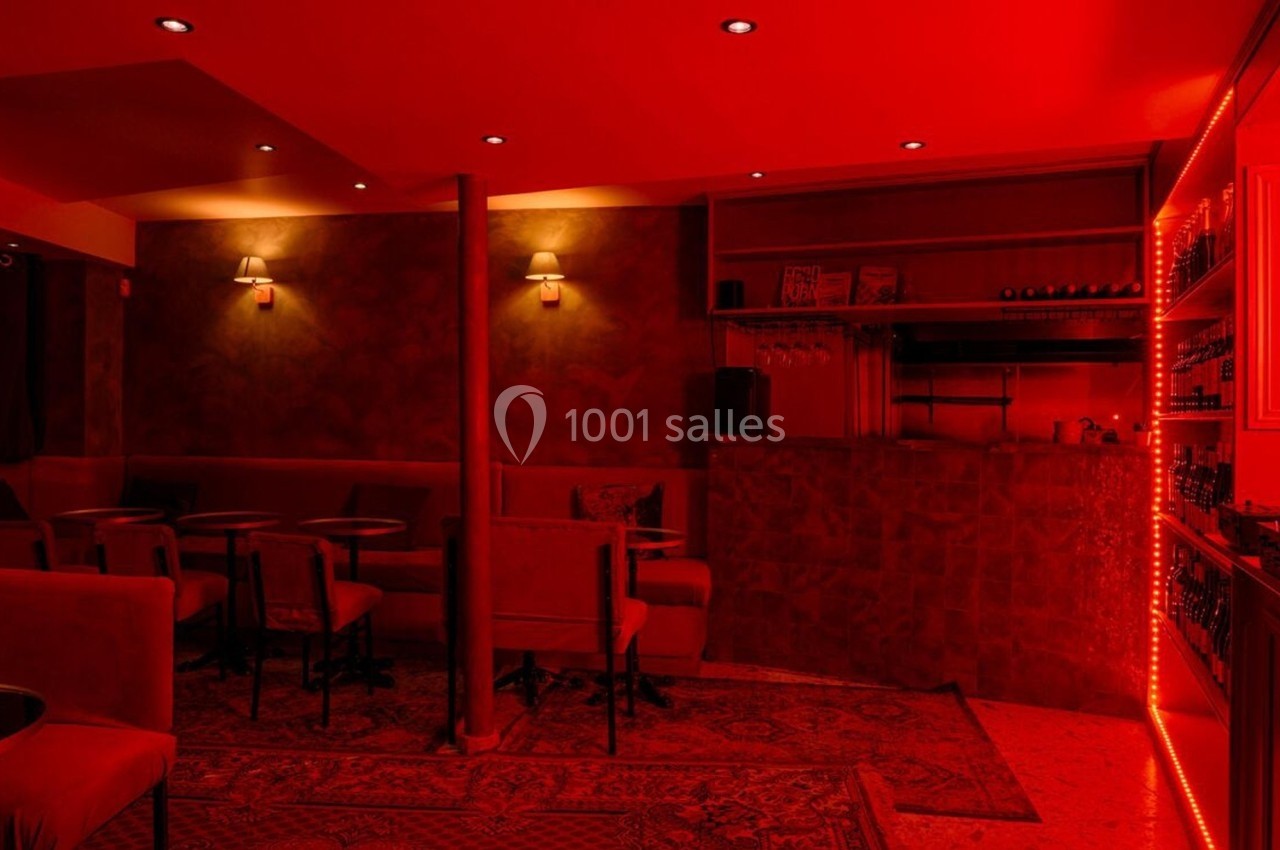 Salle de bar à l'éclairage rouge avec tables, chaises, tapis et un comptoir en arrière-plan.