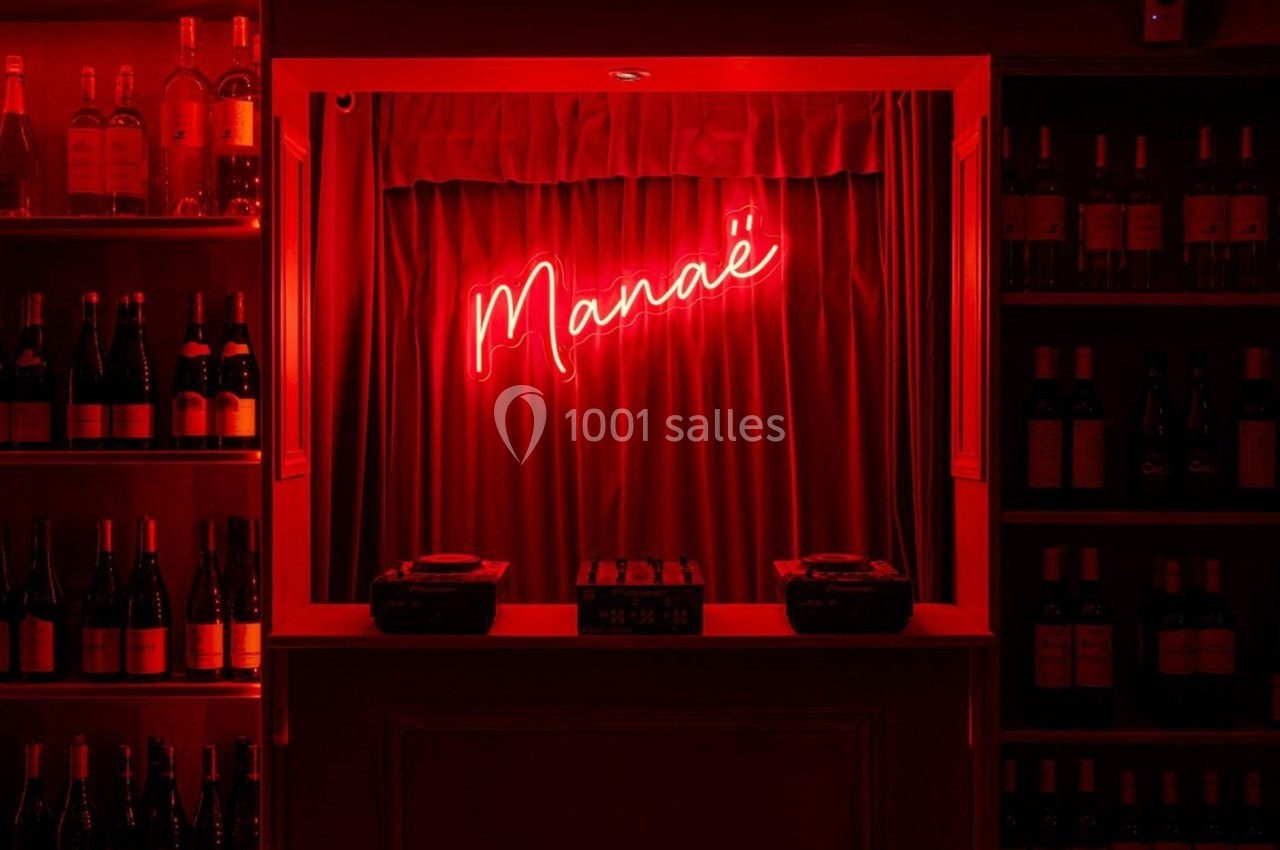 Étalage de bouteilles de vin entourant un comptoir éclairé par une enseigne néon rouge indiquant ’Manaé’.