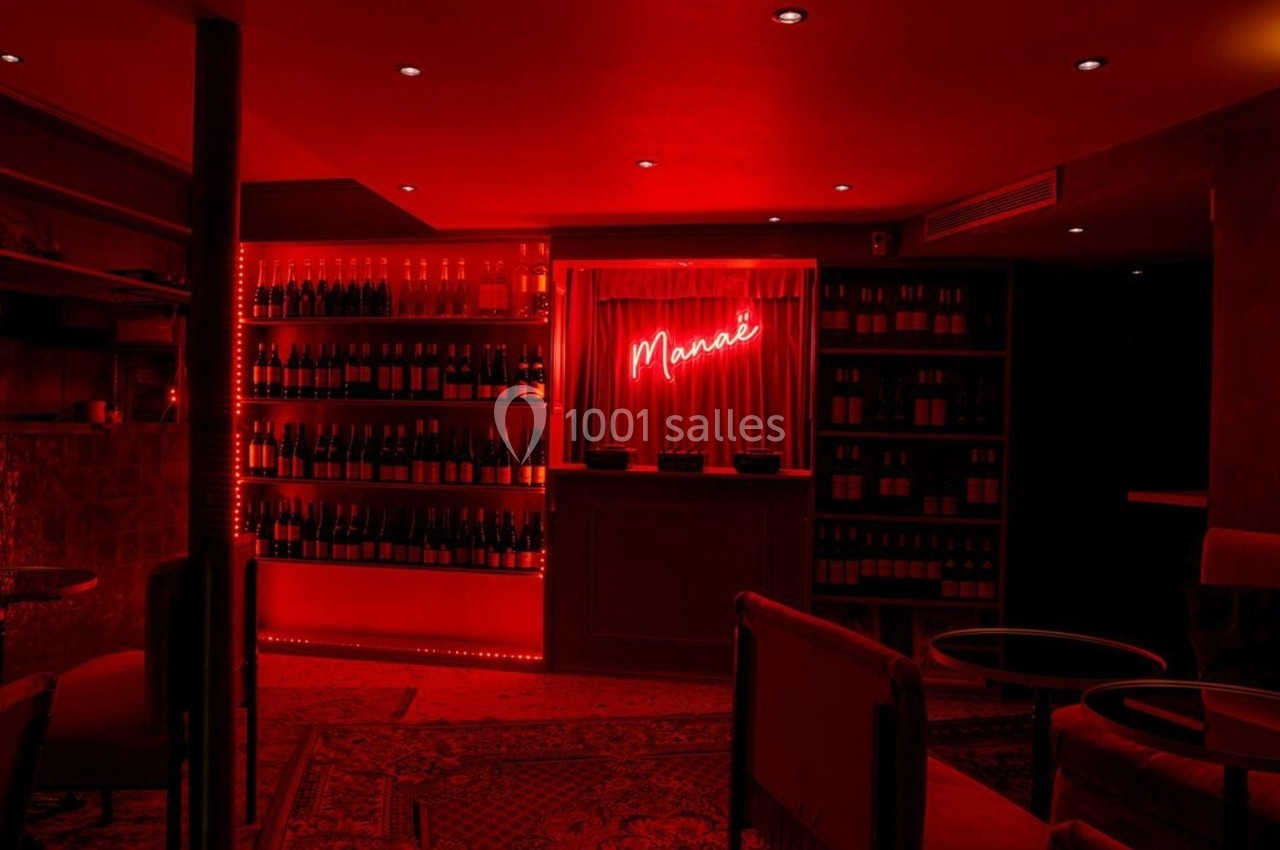 Salle tamisée avec éclairage rouge, étagères remplies de bouteilles de vin et enseigne néon « Mannaé » au mur.