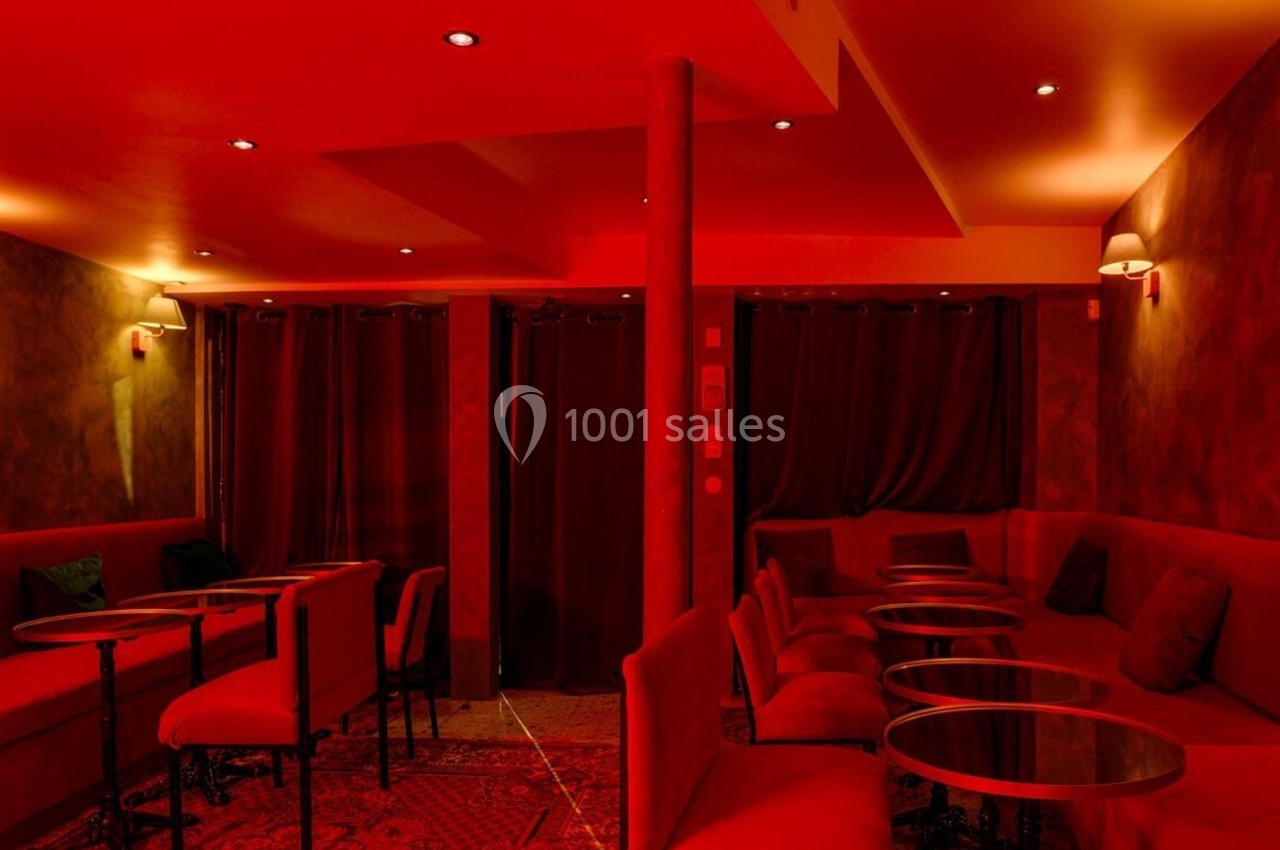 Salle intimiste éclairée de lumières rouges, avec canapés, petites tables rondes et rideaux sombres en arrière-plan.