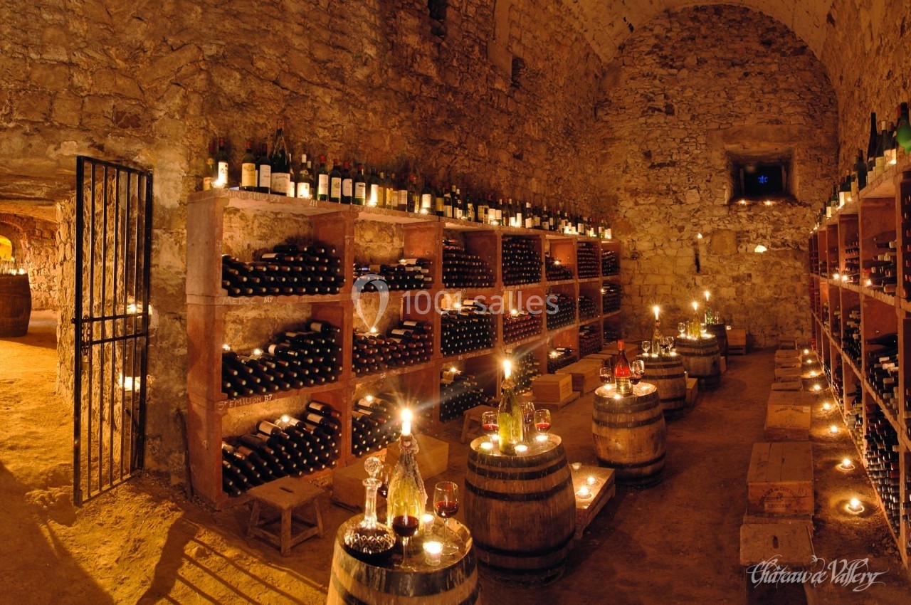 Cave voûtée en pierre avec étagères remplies de bouteilles de vin, éclairée par des bougies sur des tonneaux.