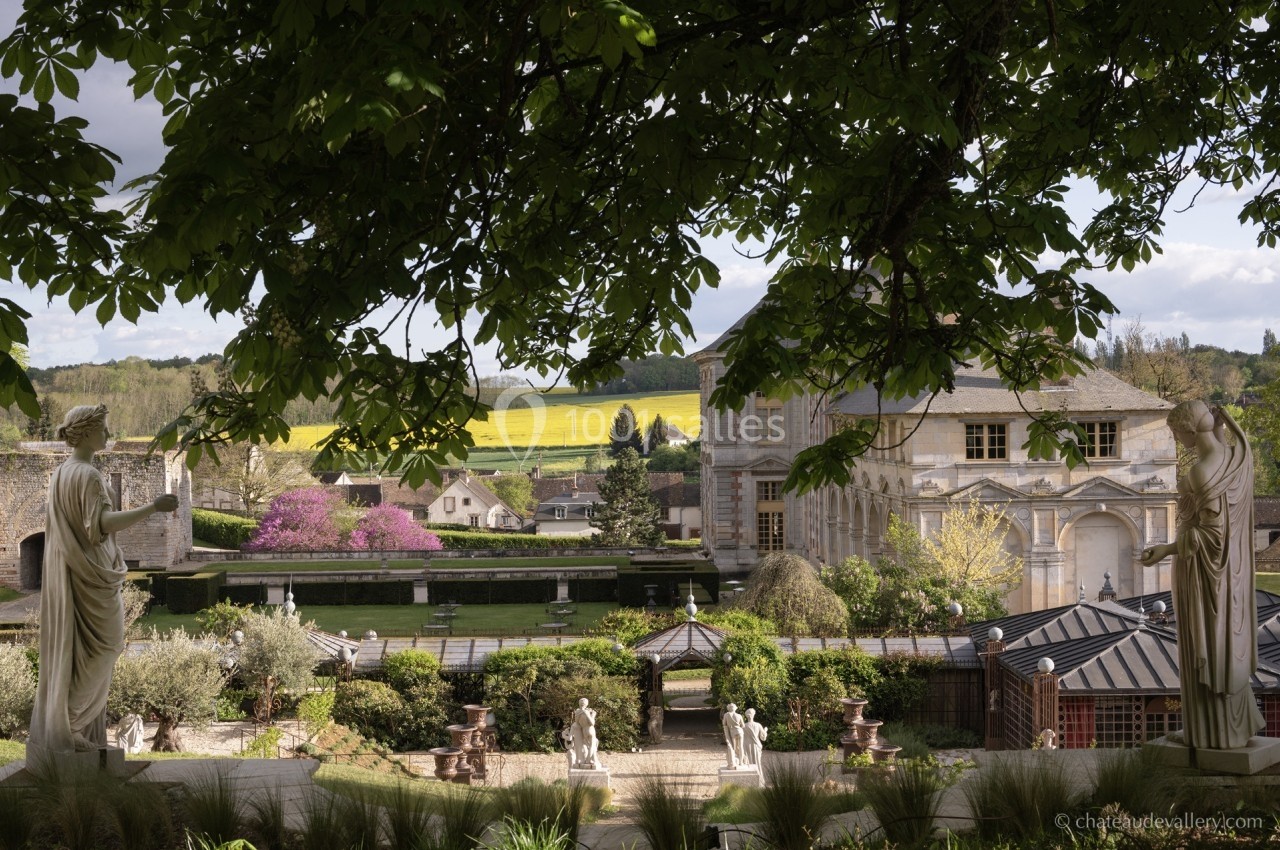 Vue d'un jardin paysager avec statues, bâtiments historiques et collines verdoyantes en arrière-plan.