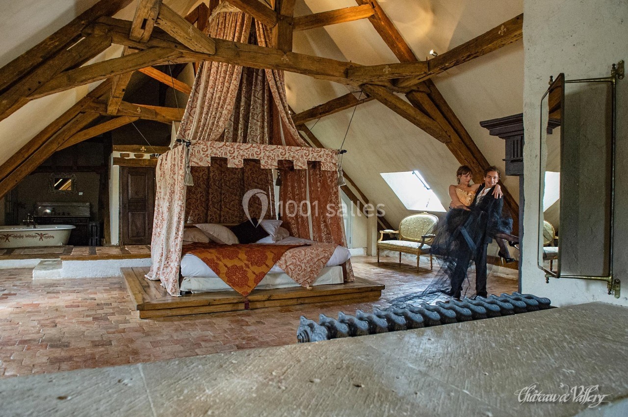 Chambre rustique avec lit à baldaquin, poutres apparentes et couple debout près d'une fenêtre.