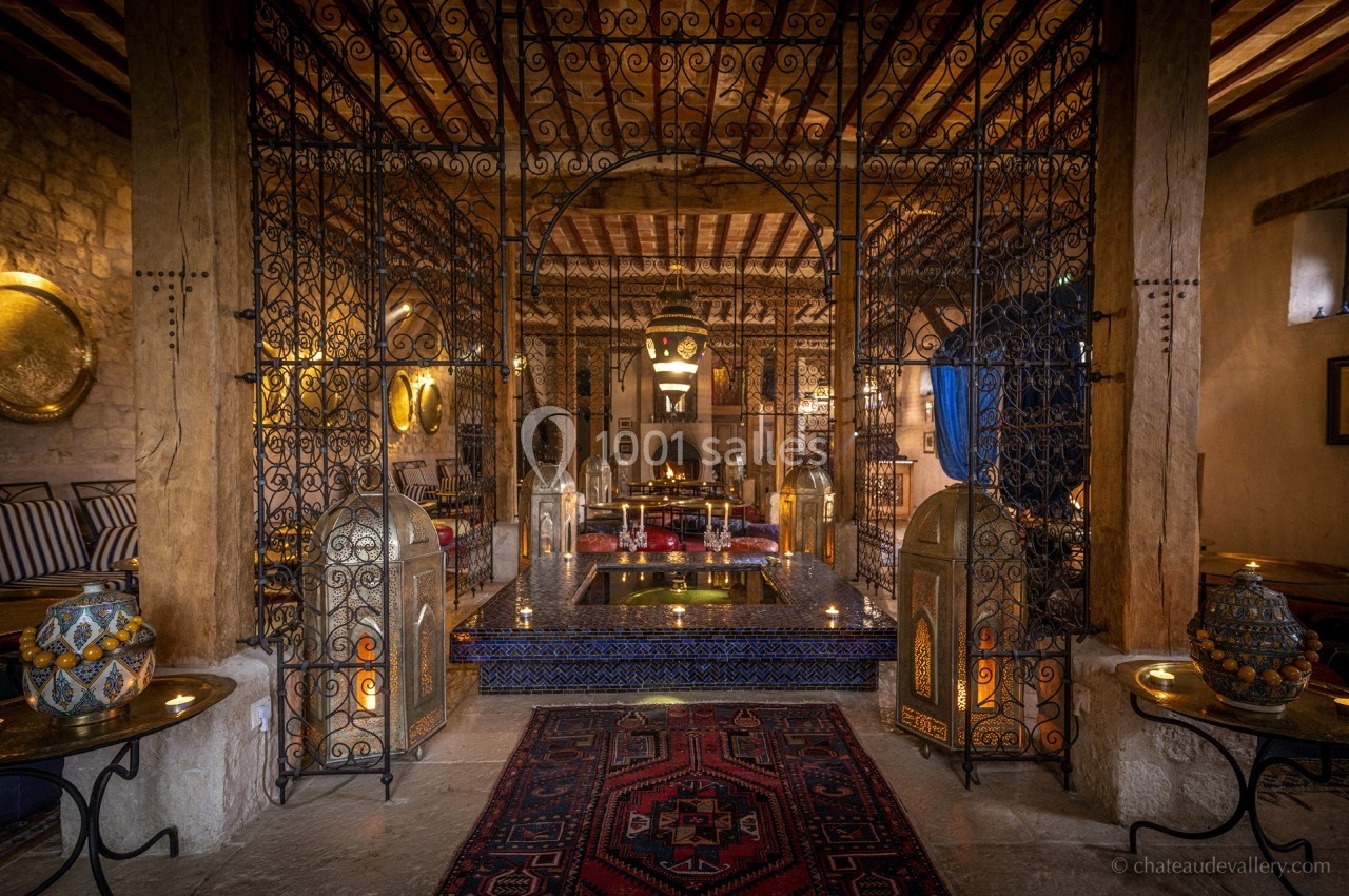 Intérieur d'une salle voûtée avec décorations orientales, lanternes, tapis et bassin central entouré de grilles en fer forgé.