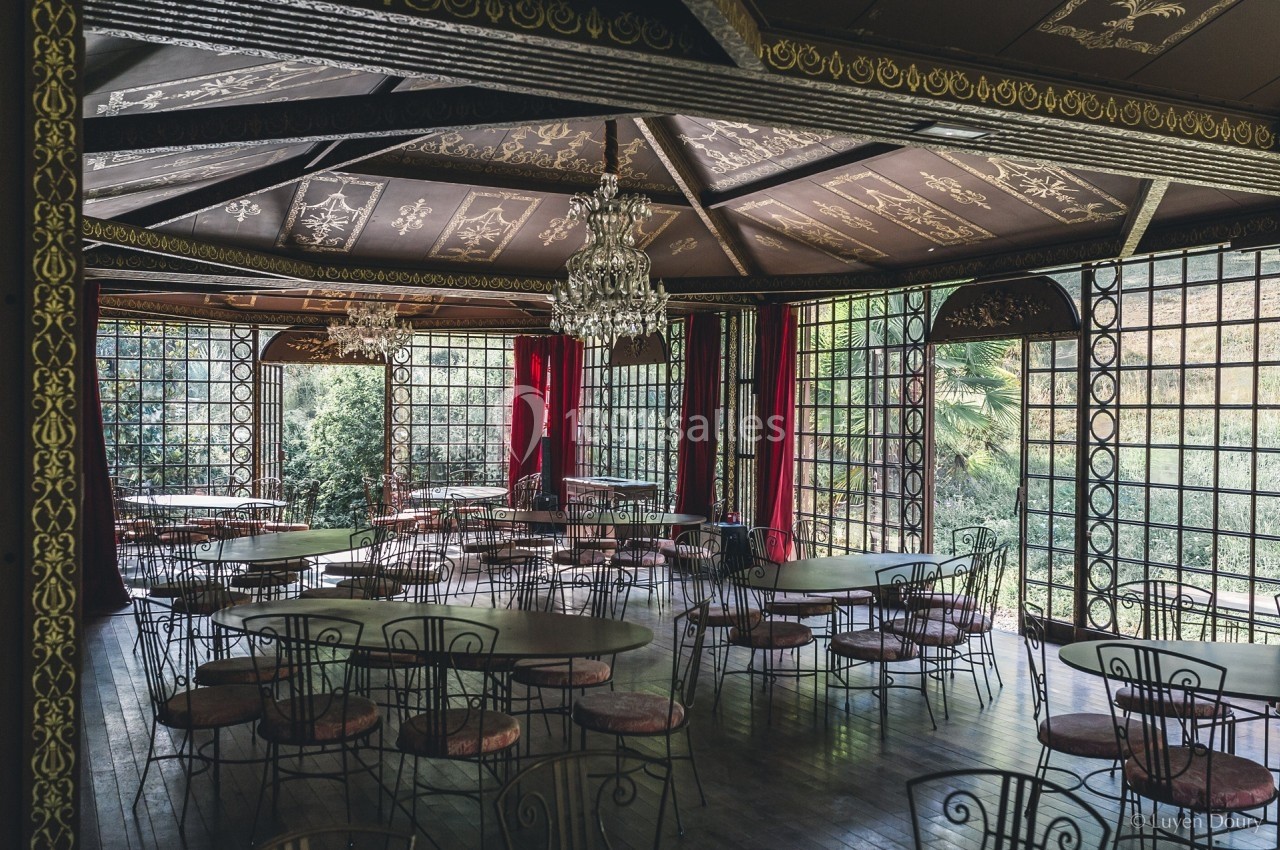 Salle lumineuse avec grandes baies vitrées, tables rondes en métal et lustres suspendus sous un plafond décoré.