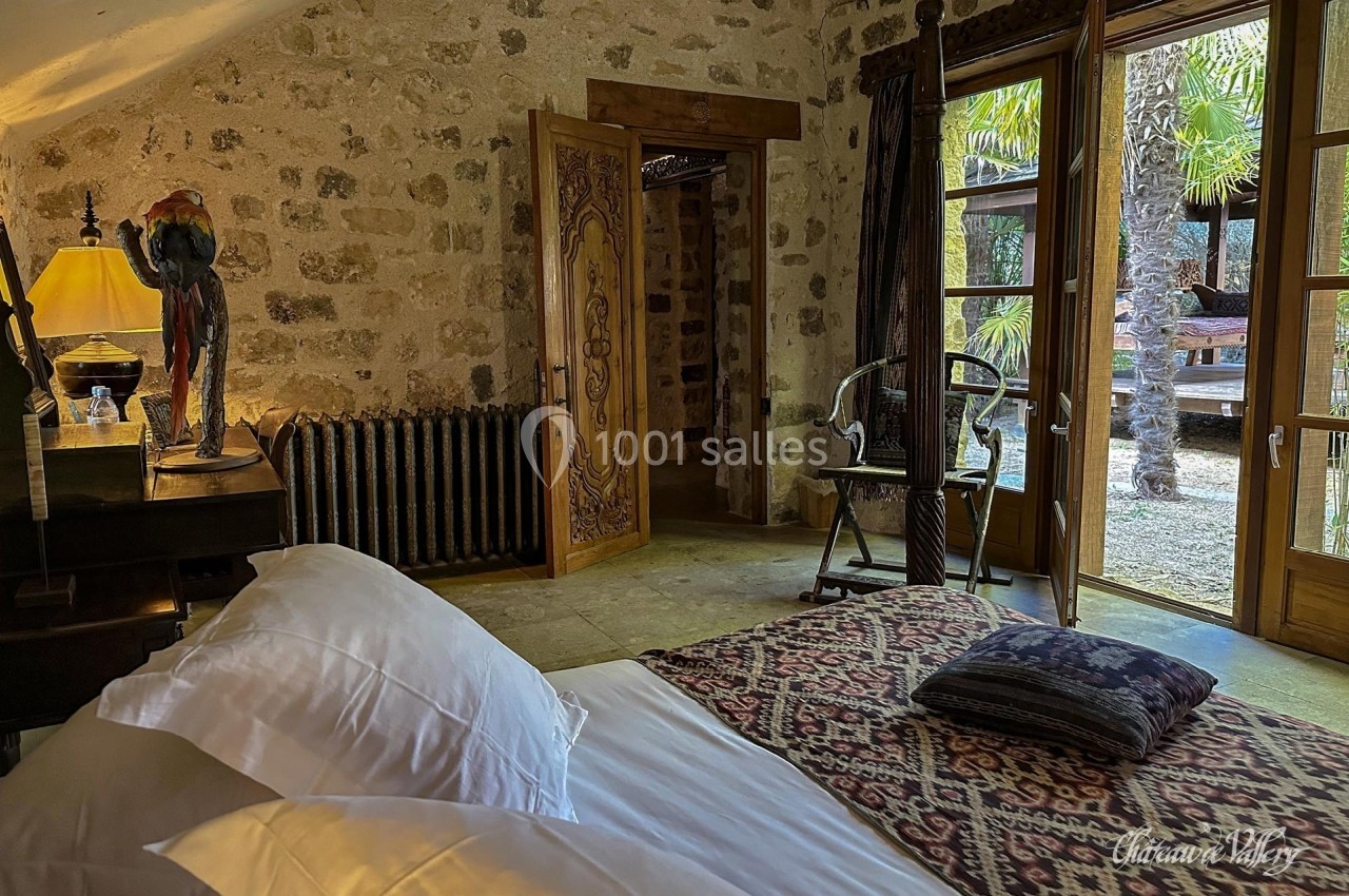 Chambre avec lit, tapis coloré, meubles en bois, et vue sur un jardin avec palmiers à travers des portes vitrées.