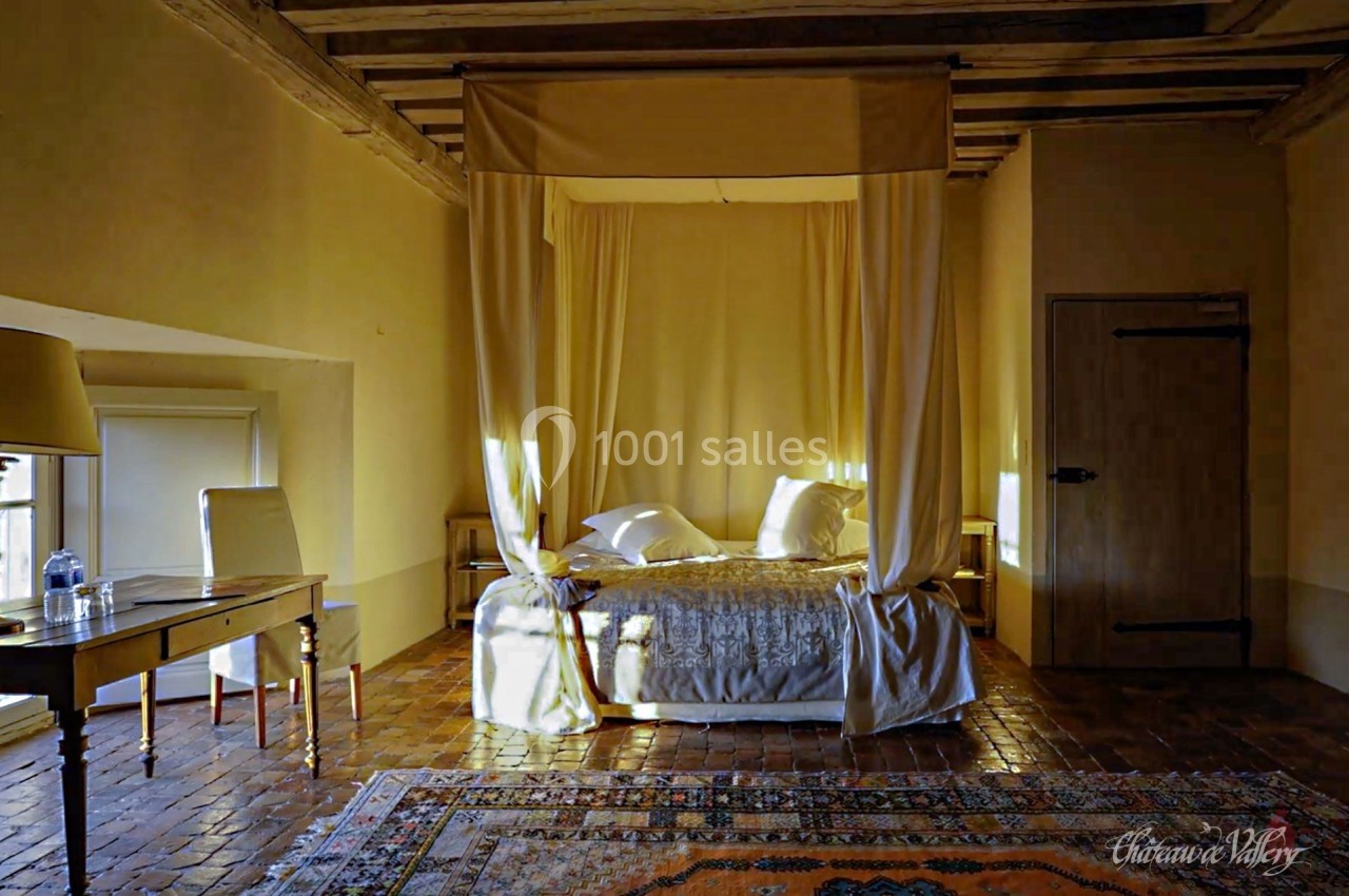 Lit à baldaquin dans une chambre rustique avec murs en pierre, parquet en bois, tapis coloré et lumière naturelle.
