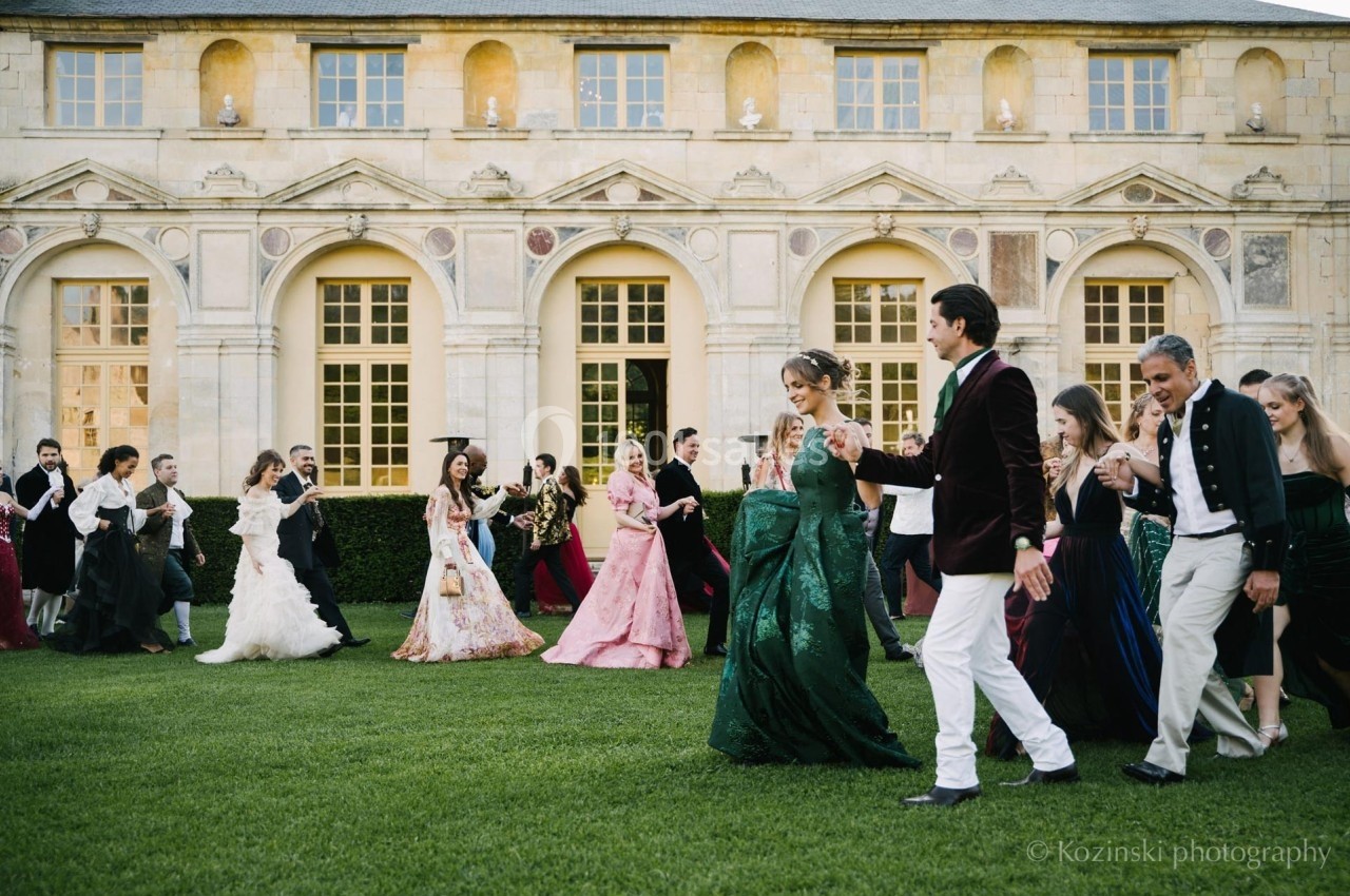 Un groupe de personnes en tenues élégantes marche sur une pelouse devant un bâtiment historique.