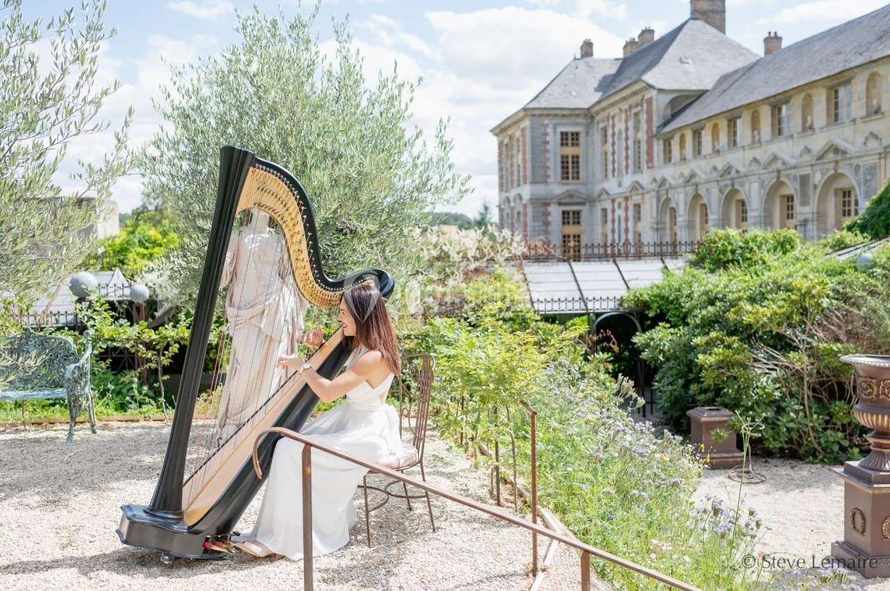 Une harpiste joue dans un jardin verdoyant, avec un bâtiment historique en arrière-plan.