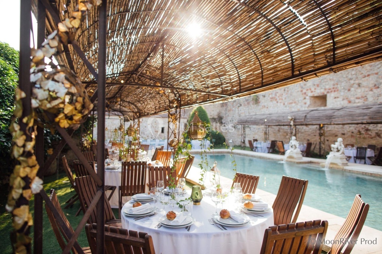 Tables dressées avec élégance sous une pergola en bambou, près d'une piscine dans une cour ensoleillée.