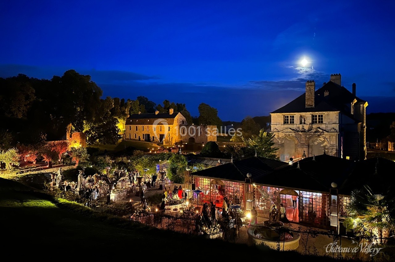 Vue nocturne d'un château éclairé avec une cour animée, des invités et une pleine lune en arrière-plan.