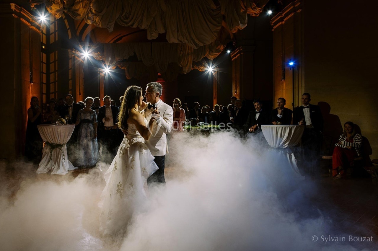 Un couple danse au milieu d'une salle éclairée, entouré de fumée et d'invités en tenue de soirée.