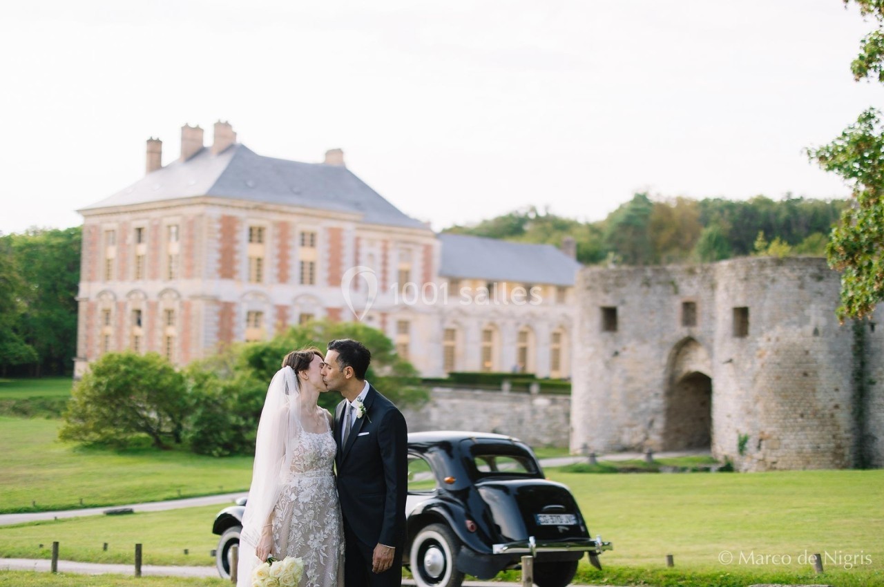 Un couple de mariés s'embrasse devant une voiture ancienne, avec un château et des ruines en arrière-plan.