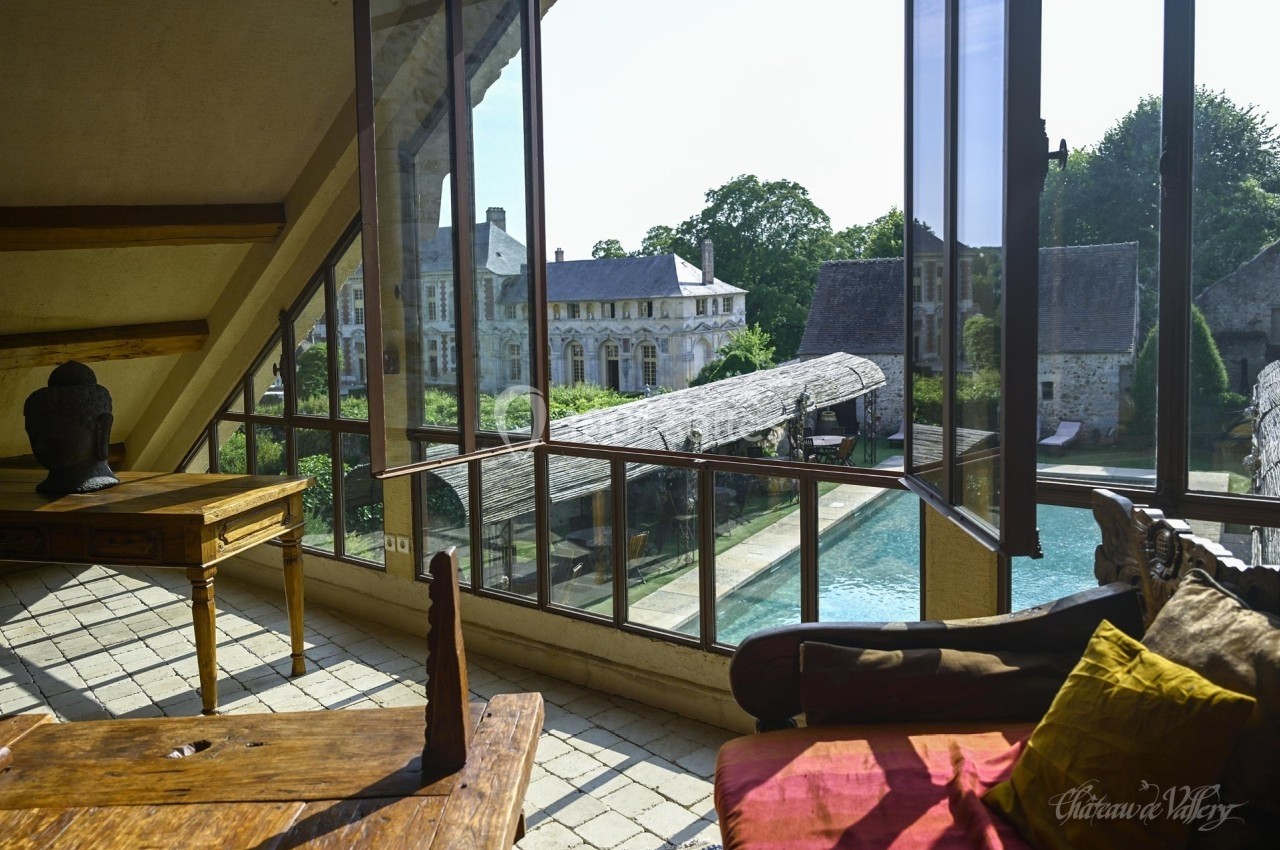 Vue depuis une pièce lumineuse avec mobilier ancien, donnant sur une piscine et des bâtiments historiques entourés de…