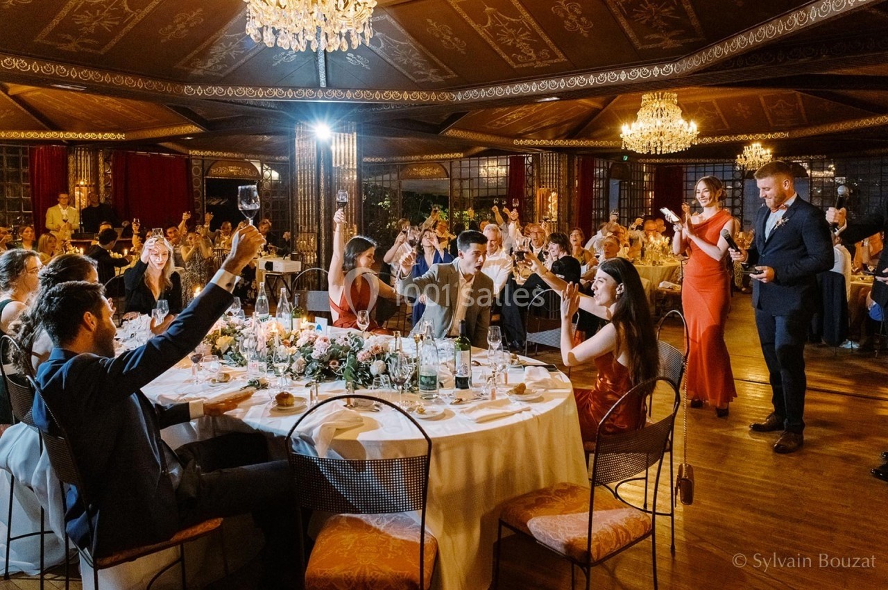 Des convives élégamment vêtus trinquent autour de tables décorées dans une salle ornée de lustres.