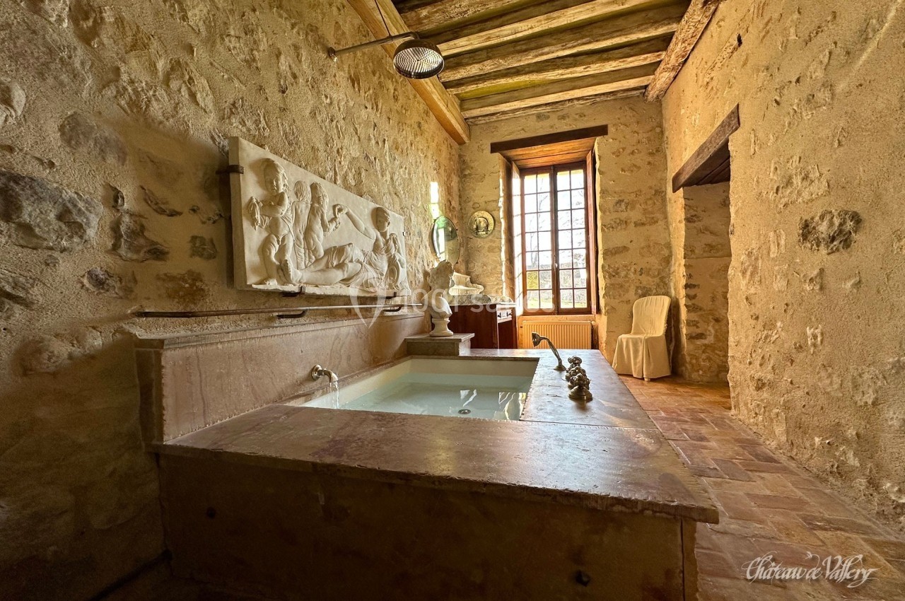 Salle de bain rustique avec murs en pierre, baignoire en pierre sculptée, fenêtre lumineuse et poutres apparentes.